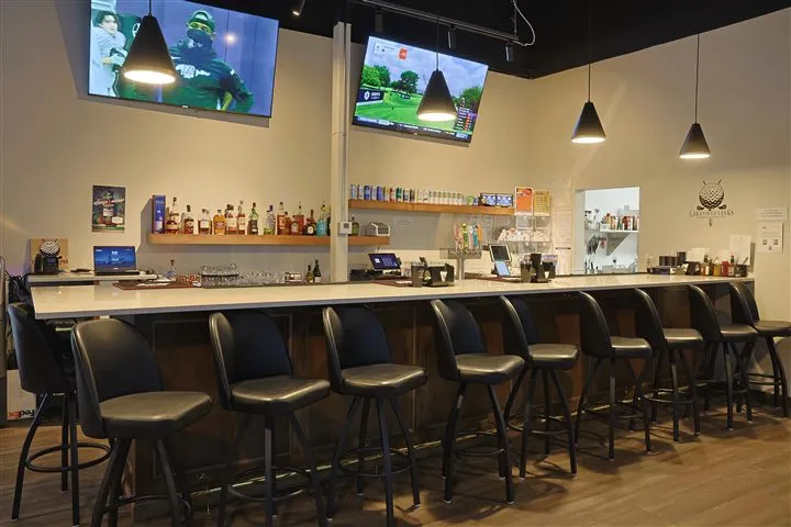 The Lakeville bar, looking down its length: a white marble-topped bar with black padded stools, shelves of liquor bottles and bar snacks against the wall, two mounted TVs showing sports and a golf broadcast, and the Lakeville Links logo on the far wall. The room is empty.