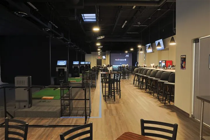 The Lakeville room seen down its length: a row of GolfZon simulator bays and putting mats under low black lighting on the left, a lit TwoVision screen at the far end, and high-top tables leading to the bar and its stools on the right, on a warm wood floor. The room is empty.