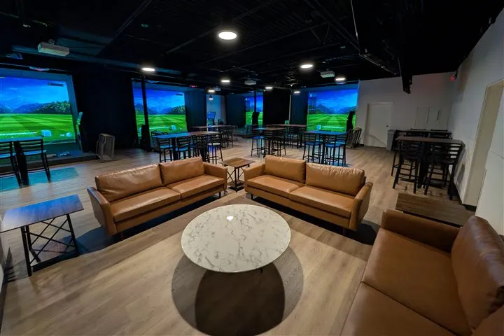 The Stillwater lounge seen from the couches: two tan leather sofas and a marble coffee table in the foreground, high-top tables behind, and three lit simulator screens along the far wall under a black ceiling hung with track lights. The room is empty.